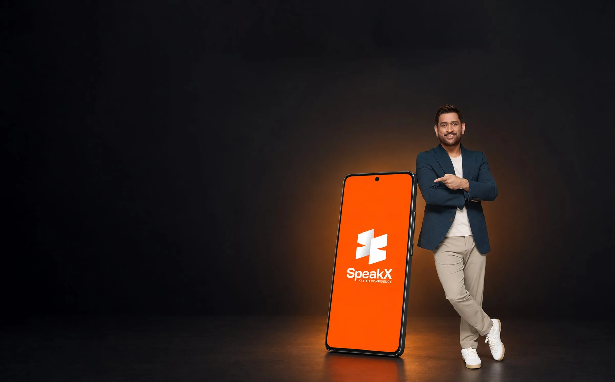 MS Dhoni in a dark studio, smiling and pointing at a phone taller than himself that he leans on; the phone shows the SpeakX splash screen.