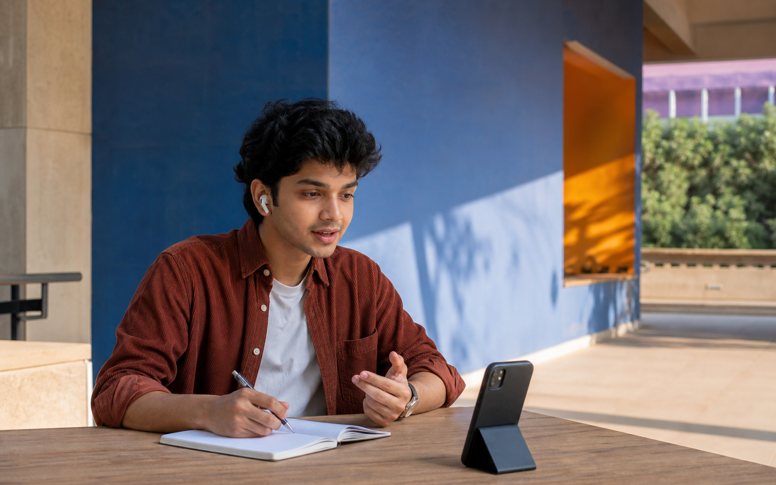 A student at an outdoor campus table practising aloud with a phone propped in front of him and a notebook open